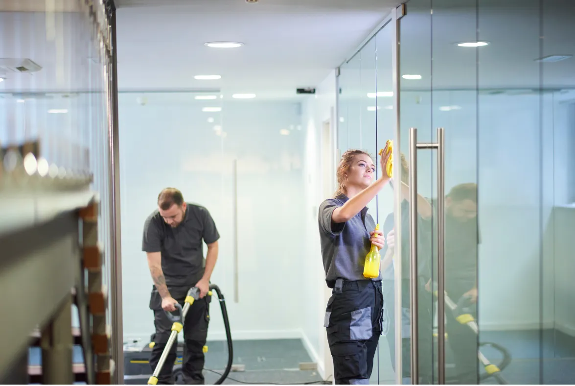 Two people cleaning a modern glass office hallway, one vacuuming and one wiping the glass door handles.